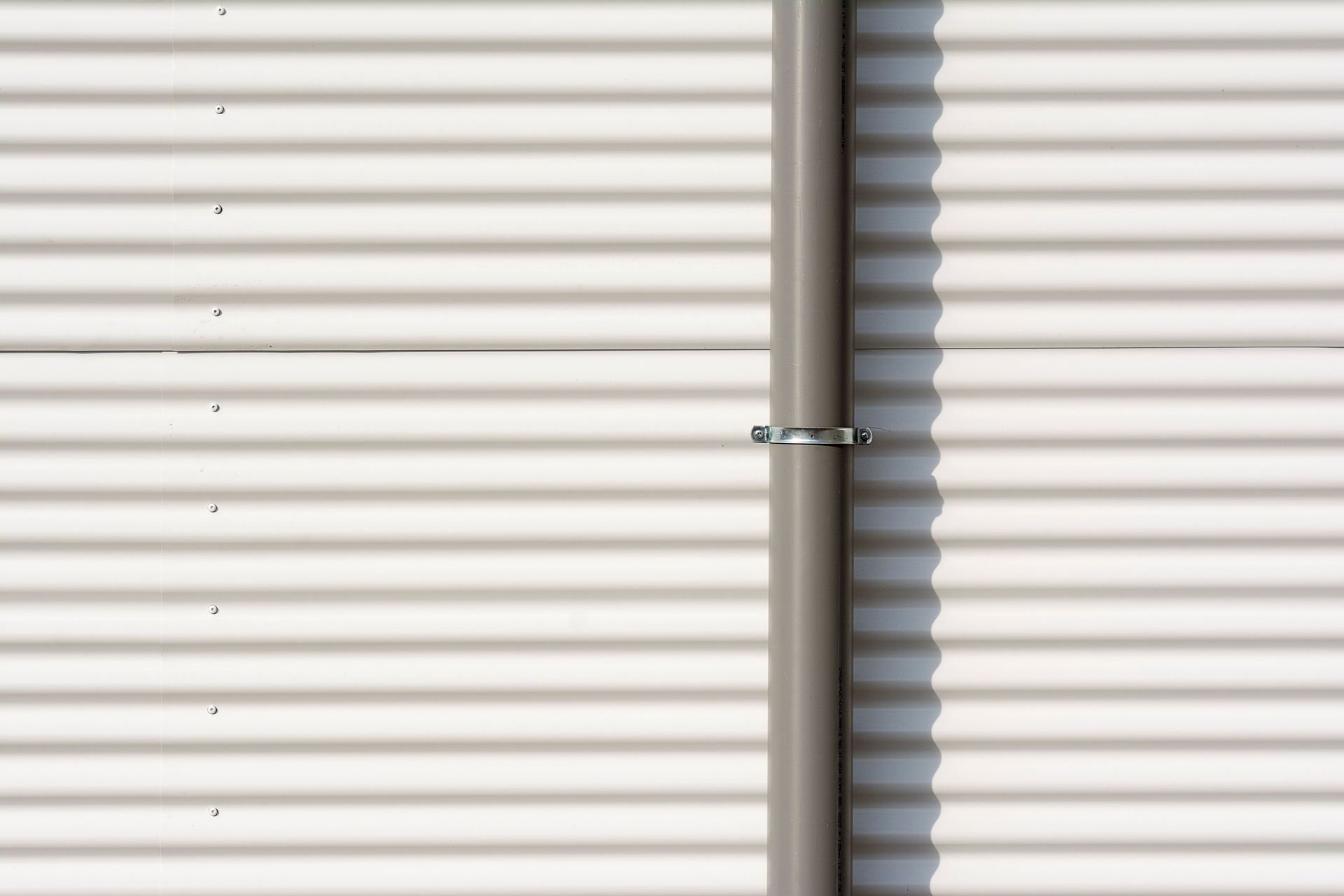 Close-up of a corrugated aluminum wall with a vertical drain pipe casting shadows.